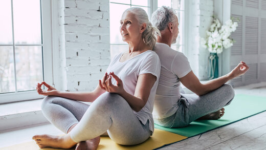 Eine ältere Damen und ein älterer Herr sitzen auf dem Fußboden und machen Yoga